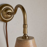 Close-up of the Vattu plug-in wall light showing the antique brass swan neck arm and round wall plate above a mango wood cone shade, with a rust-coloured cord against a plain wall.