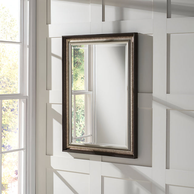 The Luxon Rectangle Wall Mirror in Antique Silver is mounted on a white paneled wall. A window beside it reflects faint sunlight and greenery, creating a serene and elegant atmosphere.