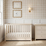 Wanderer Convertible Cot Bed with Drawer in Cashmere, cream slatted wood cot with under-bed storage drawer, shown in a nursery beside a matching dresser and gingham wallpaper.