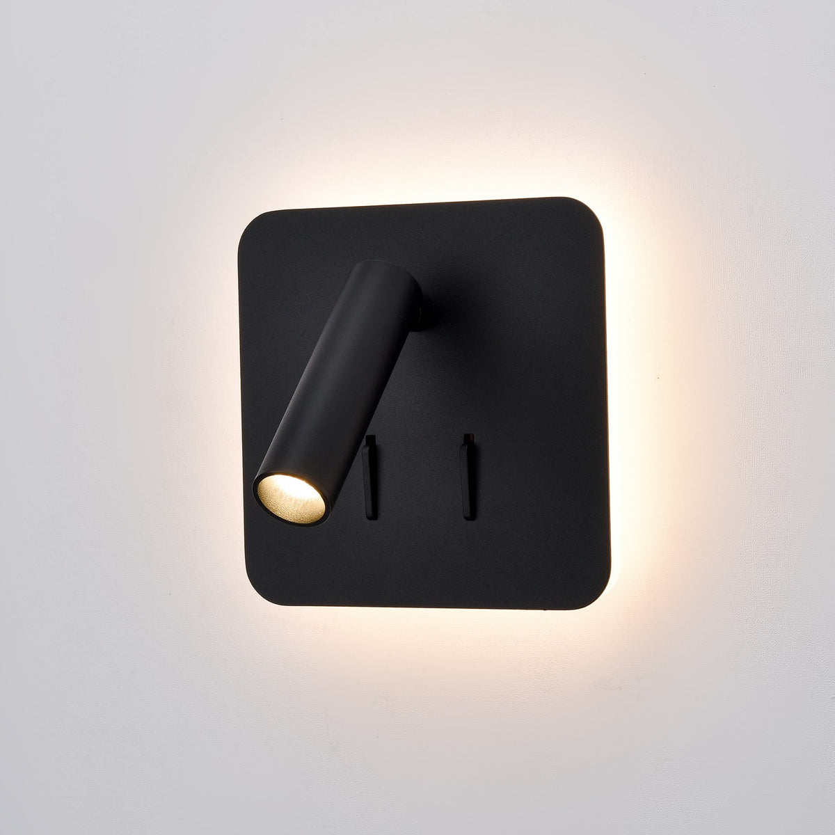 Luca Matt Black LED Wall Light with a square backplate, adjustable cylindrical reading spotlight, two toggle switches, and a soft ambient glow around the edges, shown on a plain white background.