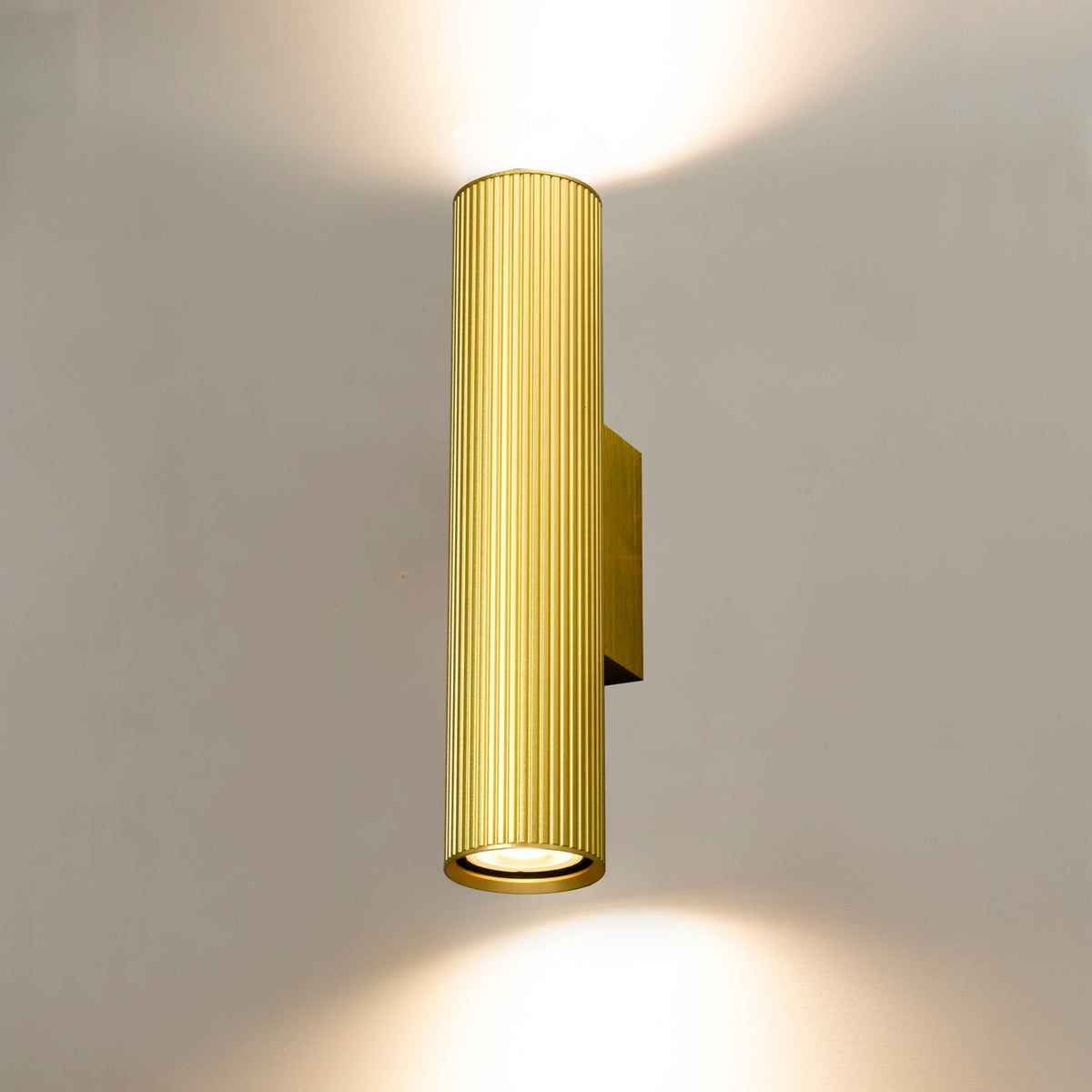 Scout Antique Brass Ribbed Wall Light with a cylindrical fluted body mounted on a square brass backplate, casting light upward and downward against a plain grey background.