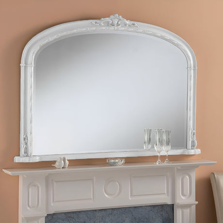 A large, ornate Brilliance Overmantle Mirror White - 130x91cm with an arched design is positioned above a white fireplace mantel. The mantel displays two glass goblets, a small dish, and ceramic figurines. The wall behind is painted in a light tan color.