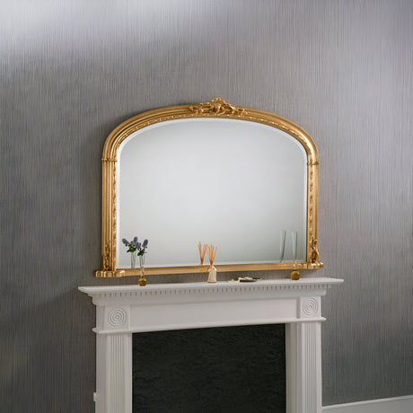 A large, ornately detailed Brilliance Overmantle Mirror Gold - 130x91cm is positioned above a white mantelpiece with decorative molding. The mantel displays a small plant, a reed diffuser, and a pair of glass vases against the backdrop of a textured gray wall.