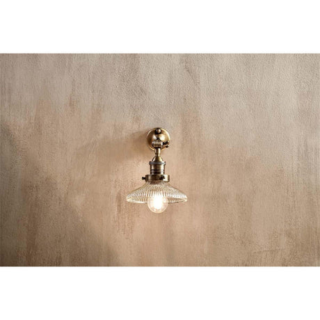 Zowie Small Recycled Glass Wall Light in antique brass, with a round wall plate, adjustable arm, and ribbed glass dish shade, shown illuminated on a plain wall.