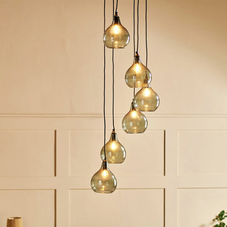 Ziva 6 Light Glass Cluster Pendant Light in Green Smoke, teardrop-shaped hammered glass shades with brass collars on black flex cords at staggered lengths, shown against a pale panelled wall.