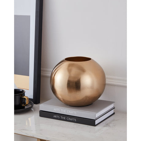 Abucay Vase 16.5cm in Gold, a round, globe-shaped aluminium vase with a brushed brass finish, shown on stacked books beside a framed print and black cup on a marble surface.