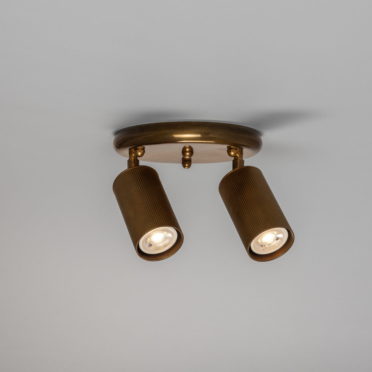 Adare Brass 2 Light Ceiling Spotlight with round ceiling plate, central ball finial, and two adjustable reeded cylindrical brass heads with GU10 bulbs, shown on a plain light grey background.