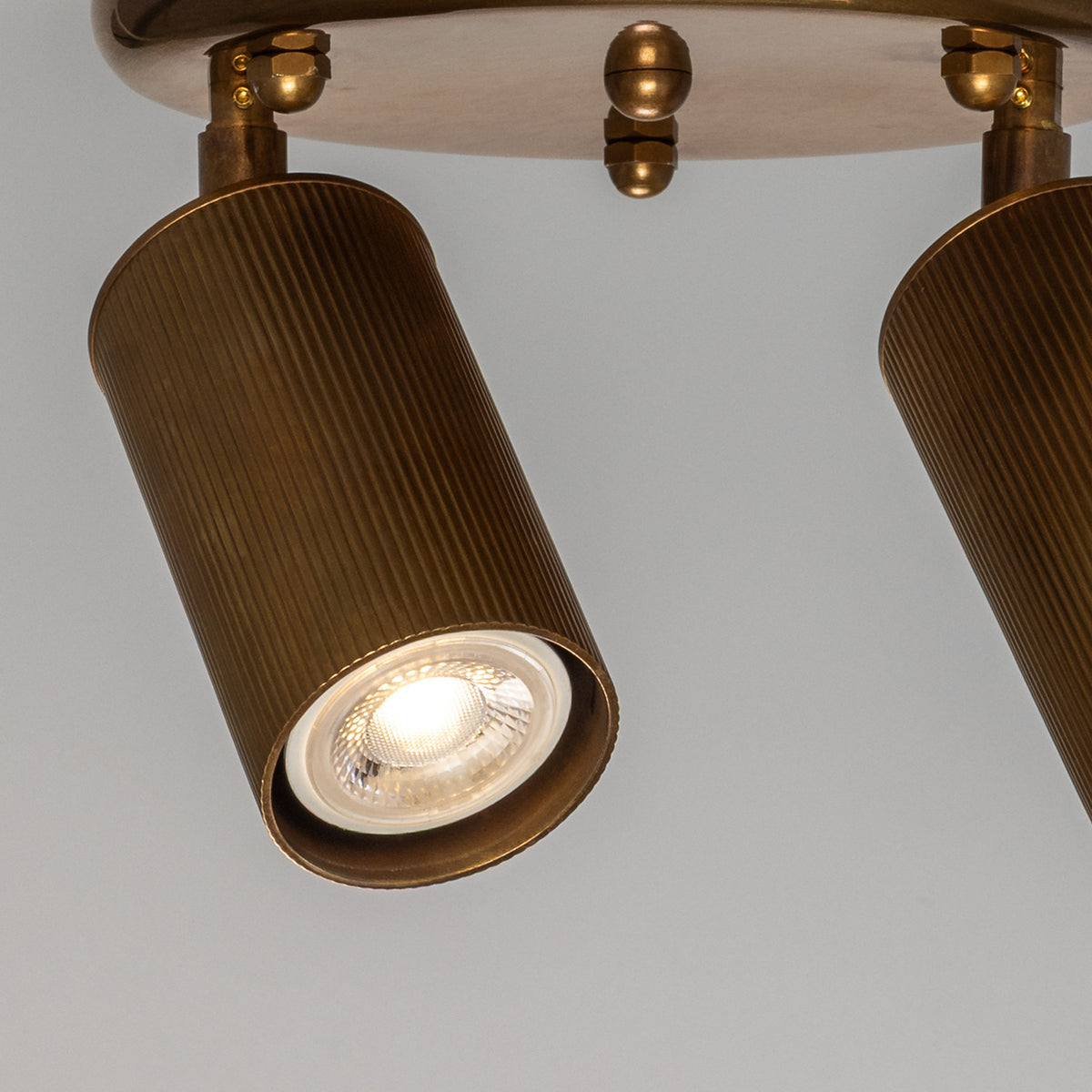 Close-up of the Adare Brass 2 Light Ceiling Spotlight, showing reeded cylindrical brass heads with GU10 bulbs on angled arms against a round ceiling plate with ball finial.