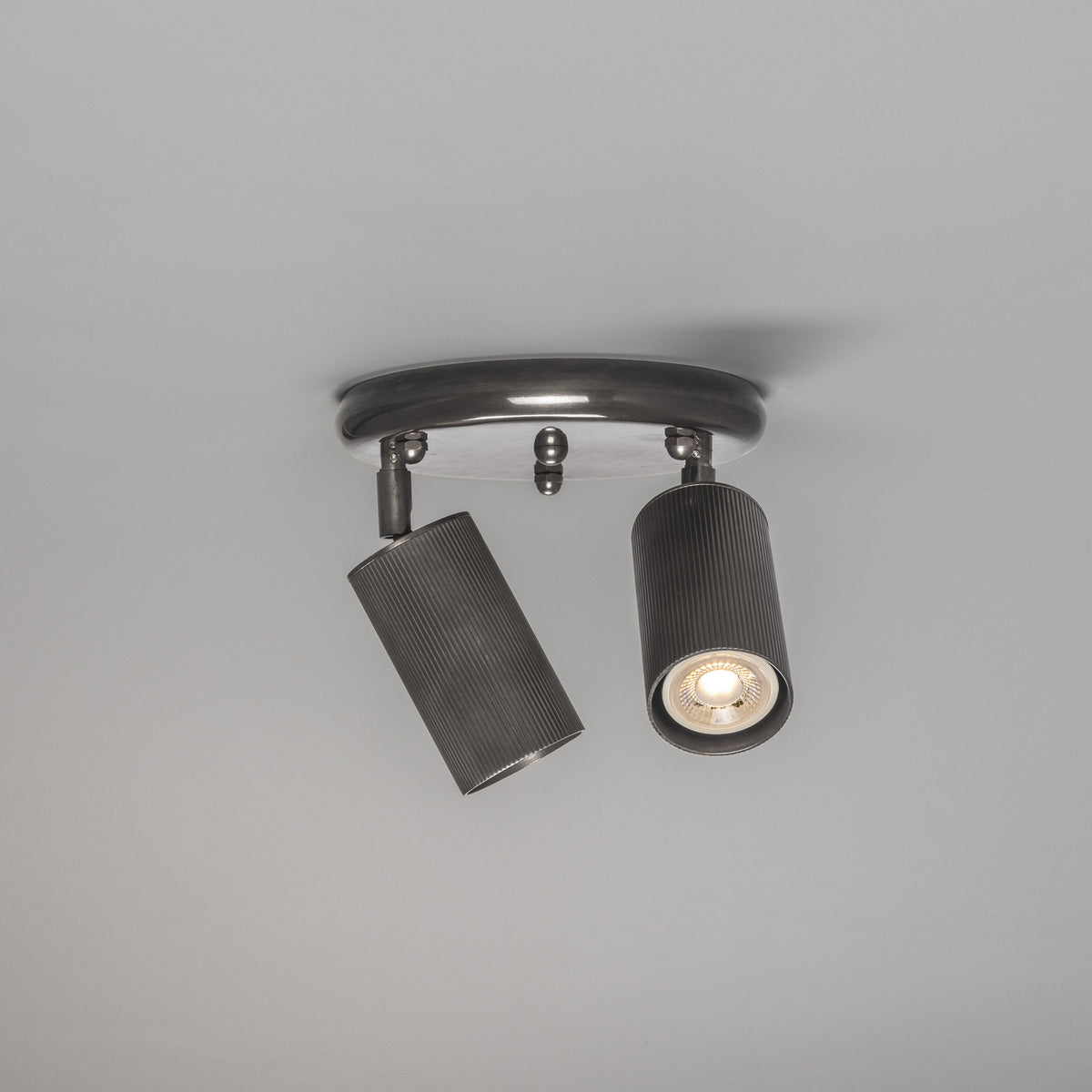 Adare Brass 2 Light Ceiling Spotlight with round ceiling plate, central ball finial, and two reeded cylindrical dark metal heads on adjustable arms, shown against a plain light grey background.