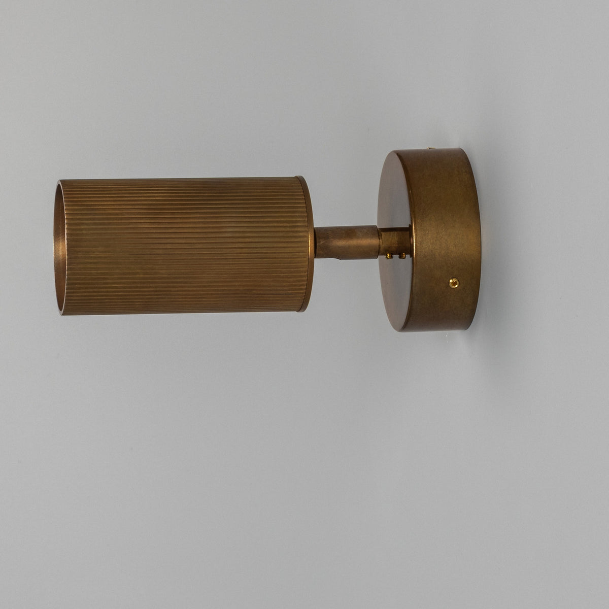 Adare Brass Wall Spotlight with ribbed cylindrical head on a short arm, mounted to a round wall plate in antique brass finish, shown against a plain light grey background.