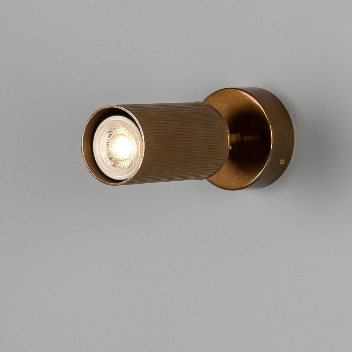 Adare Brass Wall Spotlight with ribbed cylindrical head on a short arm and round wall plate, GU10 bulb lit, shown against a plain grey background.