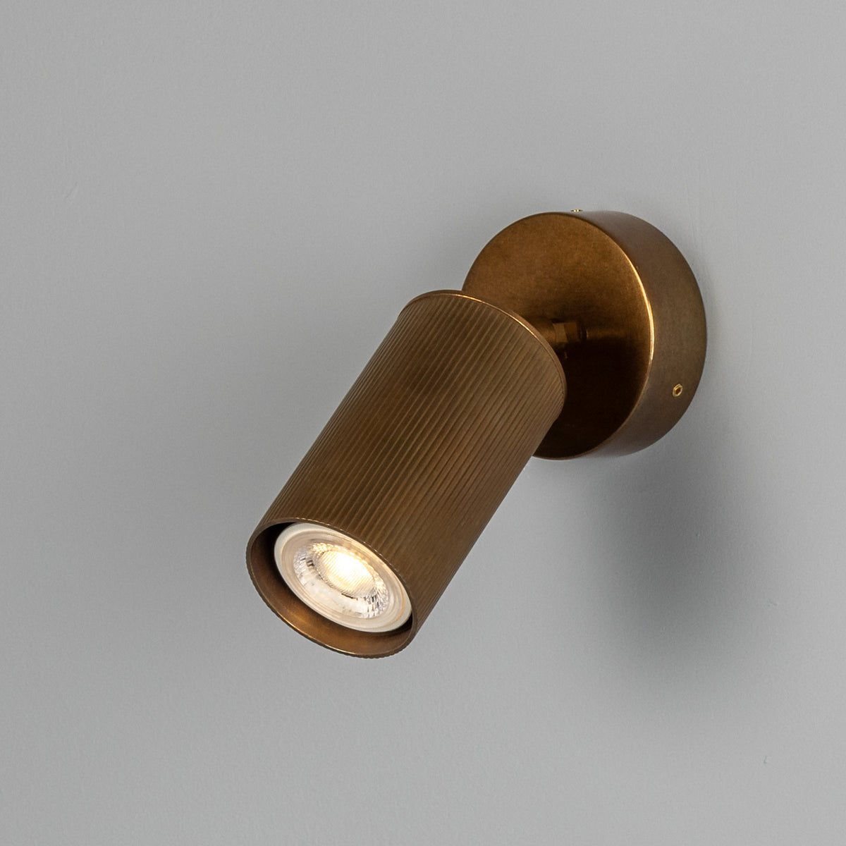 Adare Brass Wall Spotlight with ribbed cylindrical head on short arm and round wall plate, GU10 bulb visible, against a light grey background.