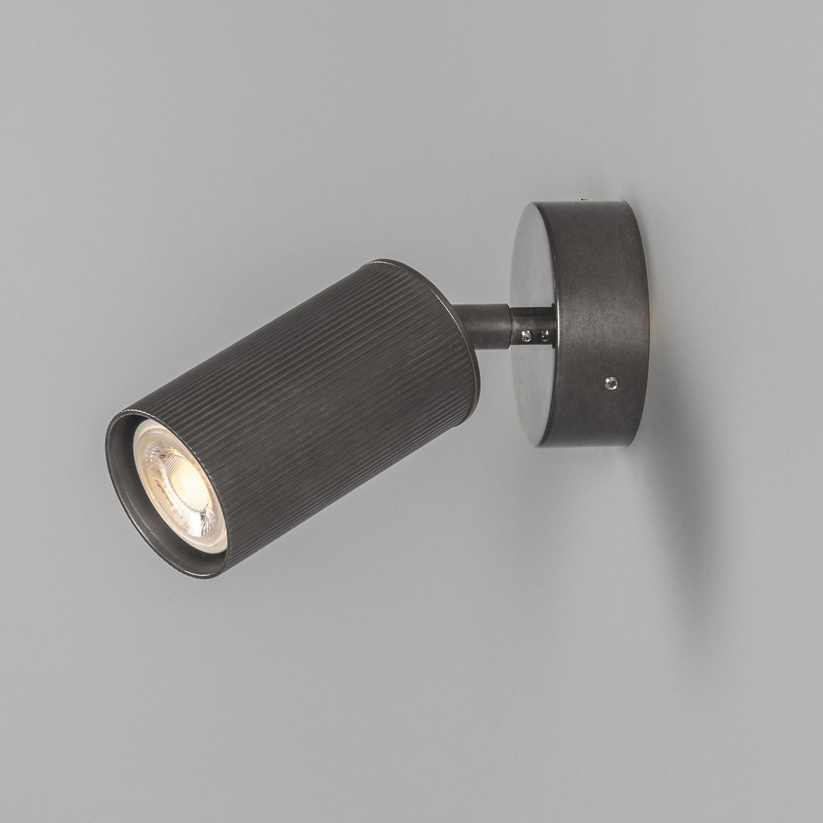 Adare Brass Wall Spotlight with a ribbed cylindrical head on a short arm and round wall plate, finished in dark antique metal, shown on a plain grey background.