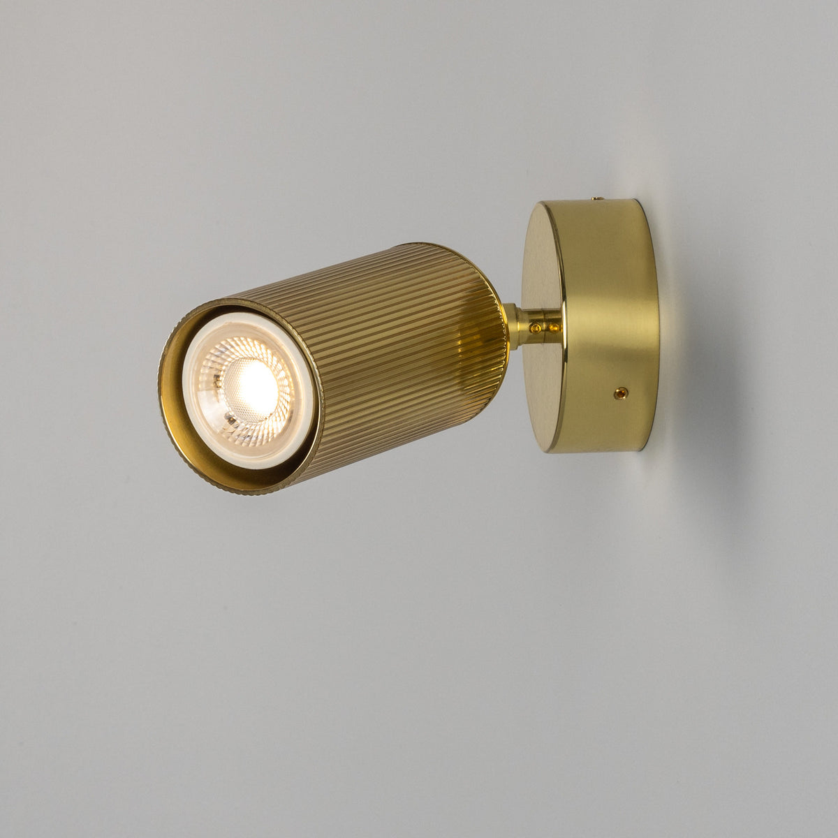 Adare Brass Wall Spotlight with ribbed cylindrical head on a short arm and round wall plate, lit GU10 bulb, shown against a plain light grey background.