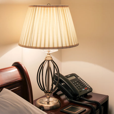 An Aegean Table Lamp - Aged Brass, warmly lit, rests on a wooden nightstand next to a telephone and smartphone. The lamp showcases a pleated shade and decorative hand-forged scrolls on its metallic base. Partial views of the edge of a bed with a pillow are in sight.