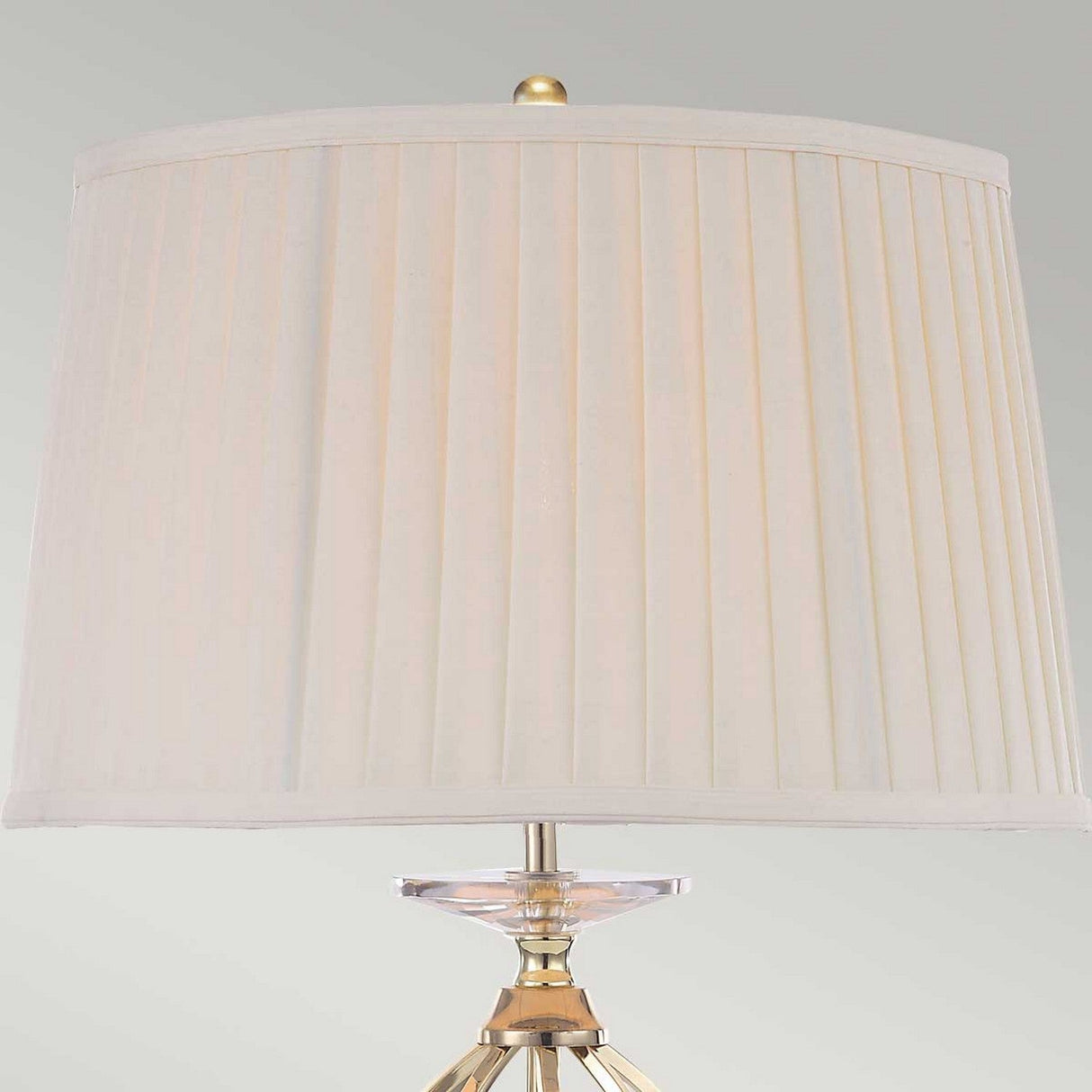 The Aegean Table Lamp - Polished Brass features a polished brass finish paired with a cream-colored, pleated lampshade set against a neutral backdrop. This elegant piece is enhanced by a clear, faceted accent located just below the shade and is complemented by its hand-forged scroll base for an extra touch of sophistication.