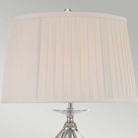 The Aegean Table Lamp - Polished Nickel showcases a pleated beige lampshade resting on a transparent, decorative base, with its polished nickel accents standing elegantly against a plain gray wall.