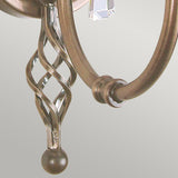 Close-up of an ornate, aged brass lamp arm featuring an intricate twisted design and a pendulum-like end, accentuated by a faceted crystal hanging beneath it. The soft gray background evokes the elegance associated with the Aegean Wall Light in Aged Brass.