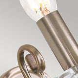 A close-up of an Aegean Wall Light - Aged Brass shows an aged brass sconce with a spiral arm and exposed bulb, set against a light gray background. The metal's brushed finish accentuates its classic elegance.