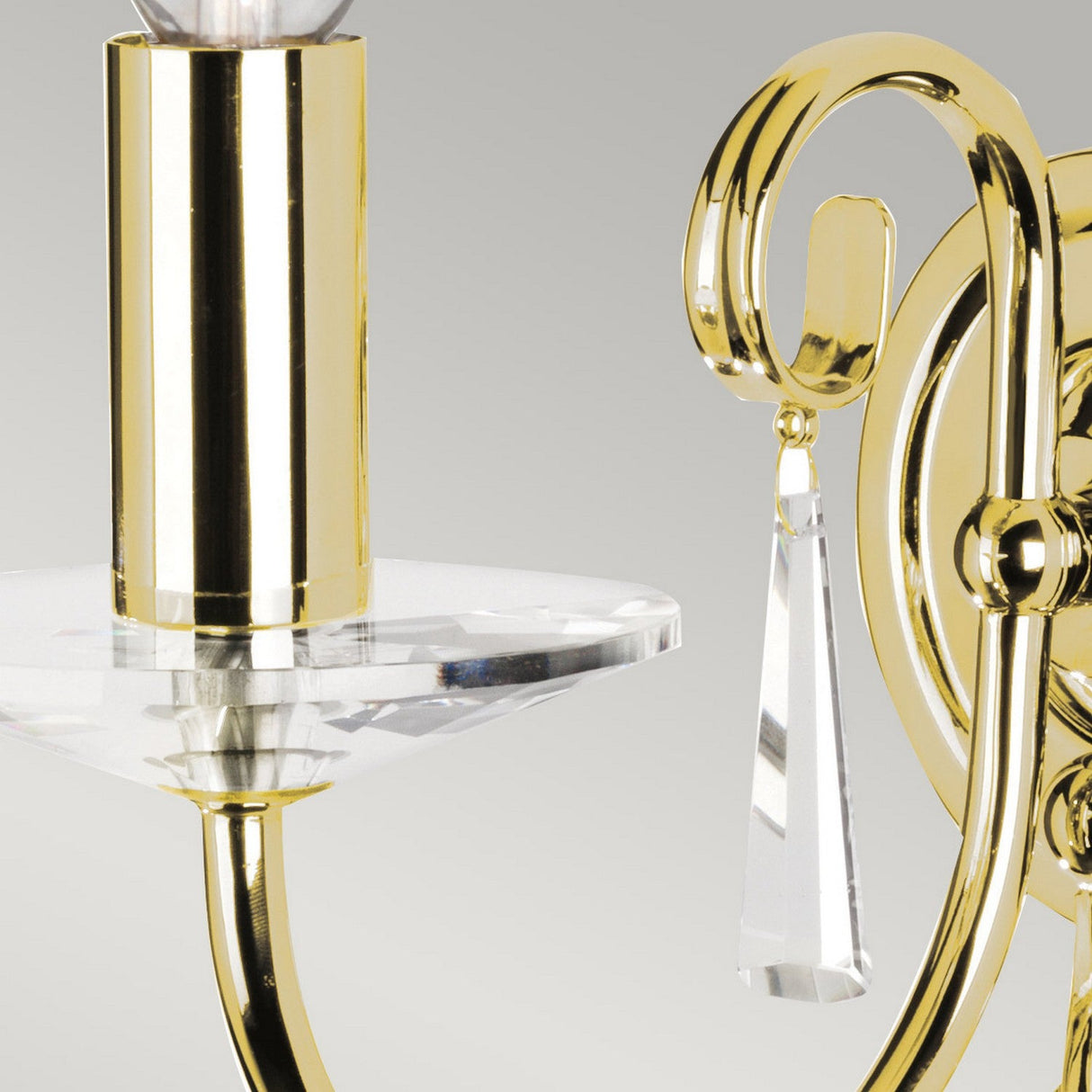 Close-up of the Aegean Wall Light - Polished Brass showcasing an elegant gold sconce with a candle-style fixture, embellished with hand-forged scrolls and a crystal pendant. This stunning piece is mounted on a round base, offering a striking polished brass design against the neutral gray backdrop.