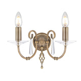 Presenting the Aegean 2 Light Wall Light in aged brass, featuring an antique-style design with two gracefully curved arms, each holding a candle-shaped bulb. Sparkling cut-glass droplets hang below each bulb for an exquisite touch. The circular base is enhanced by a decorative finial for added elegance.