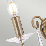 Close-up of the Aegean 2 Light Wall Light in aged brass, featuring a candle-shaped bulb and hand-forged scrolls with a decorative crystal element set against a light gray background.