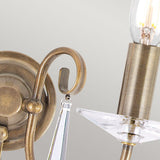 Detailed view of the Aegean 2 Light Wall Light in aged brass, showcasing its candle-style bulb. The fixture boasts a hand-forged scroll arm and decorative cut-glass droplets, set against a neutral background.