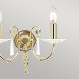 Introducing the Aegean 2 Light Wall Light in polished brass, featuring a luxurious gold sconce design with two elegantly curved arms that hold candle-shaped bulbs. Each arm is adorned with decorative crystals and hanging cut-glass droplets, all set against a subtle light gray background.