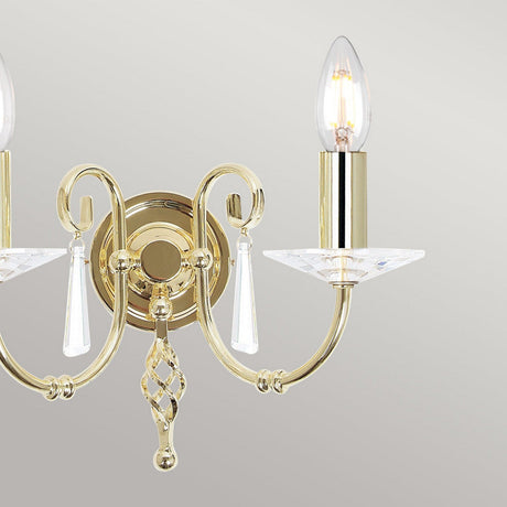 Introducing the Aegean 2 Light Wall Light in polished brass, featuring a luxurious gold sconce design with two elegantly curved arms that hold candle-shaped bulbs. Each arm is adorned with decorative crystals and hanging cut-glass droplets, all set against a subtle light gray background.