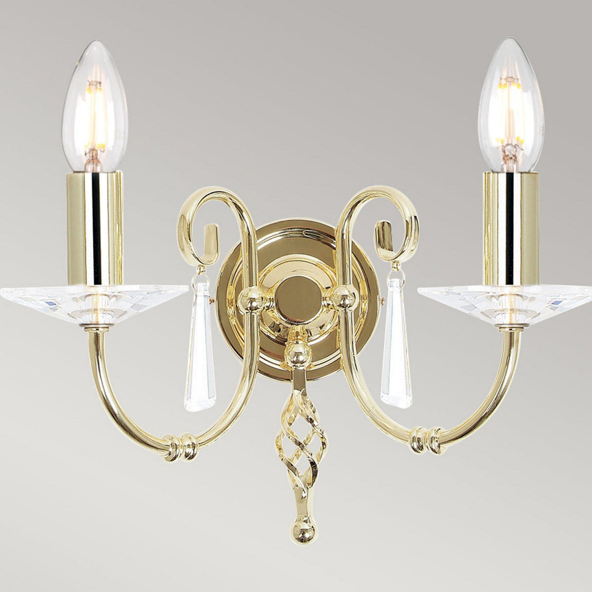 The Aegean 2 Light Wall Light in polished brass is a luxurious fixture featuring two candle-shaped bulbs. With its elegant scrollwork and cut-glass droplets on a circular base, it emits a warm glow, highlighting its classic and sophisticated design.