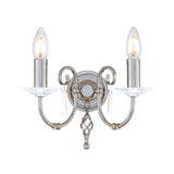 The Aegean 2 Light Wall Light in a polished nickel finish features a decorative design with two upward-facing candle-style bulbs on hand-forged scrolls, embellished with cut-glass droplets and attached to a round base.