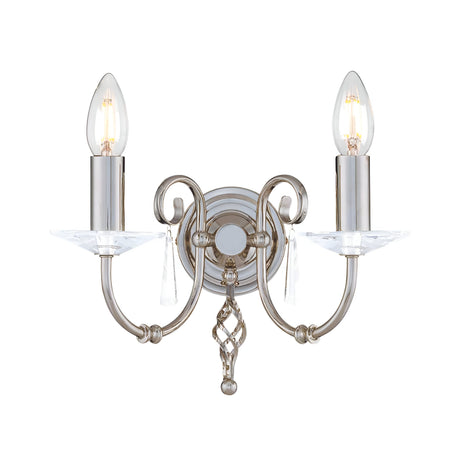 The Aegean 2 Light Wall Light in a polished nickel finish features a decorative design with two upward-facing candle-style bulbs on hand-forged scrolls, embellished with cut-glass droplets and attached to a round base.