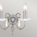 The Aegean 2 Light Wall Light in polished nickel, with two candle-style light bulbs and cut-glass droplets, hangs on a pale gray wall. This fixture showcases elegant hand-forged scrolls and dangling crystal decorations.