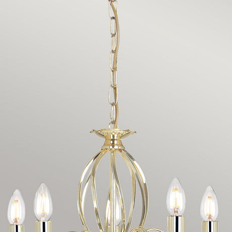 The Aegean 5 Light Chandelier in polished brass elegantly hangs from the ceiling with its decorative chain. It is handcrafted and illuminates the space with multiple candle-shaped light bulbs, set against a plain, light gray backdrop.