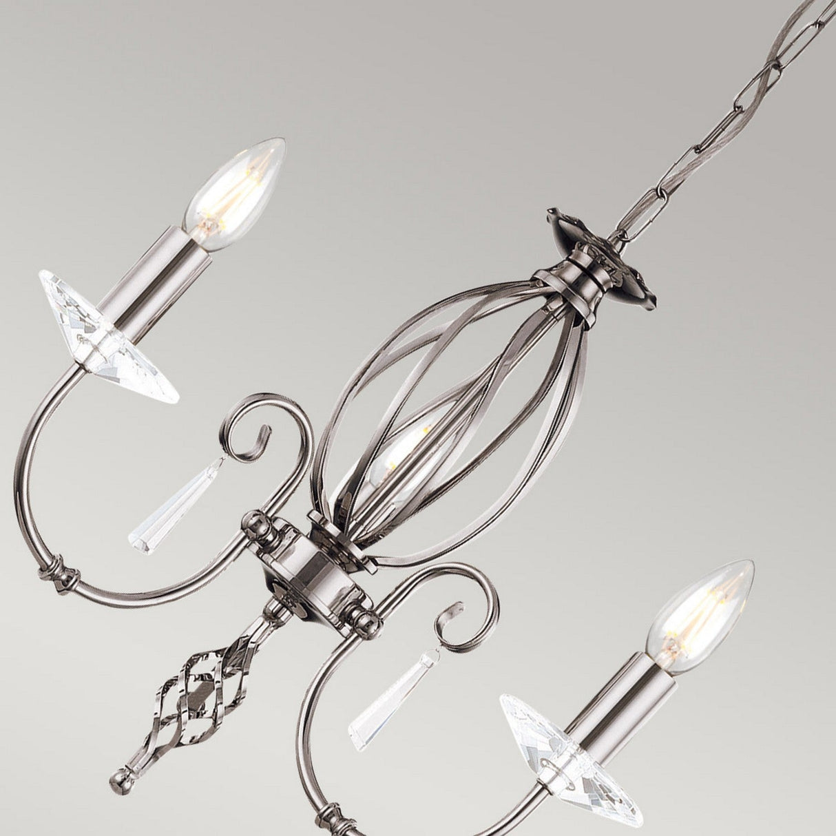 A close-up of the Aegean 5 Light Chandelier in polished nickel showcases its elegance, featuring three candle-shaped bulbs and decorative crystal accents. Hand-forged scrolls and intricate metalwork enhance its charm as it hangs against a neutral background.