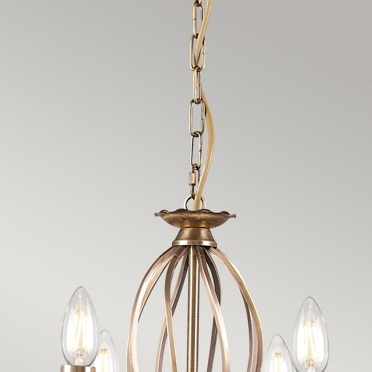 A close-up of the Aegean 8 Light Chandelier - Aged Brass highlights its hand-forged metal chain link. The chandelier showcases candle-shaped bulbs within an open, cage-like design, all set against a plain gray background.