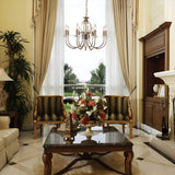 Elegant living room with tall windows, beige curtains, and the Aegean 8 Light Chandelier in aged brass, adding a touch of vintage charm. Two striped armchairs face a wooden coffee table adorned with a floral centerpiece. A large plant stands beside the chairs, and there's an ornate fireplace to the right, framed in aged brass detailing.