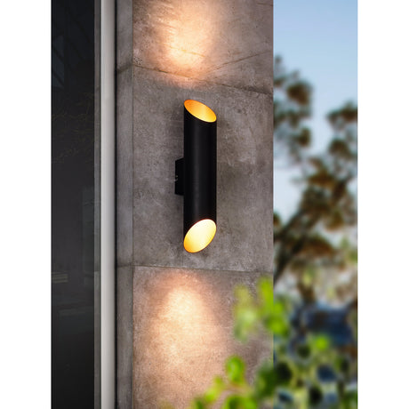 Agolada LED Outdoor Wall Light in black steel with copper interior, mounted on a stone wall, up-and-down light from angled tube ends, IP44 rated.