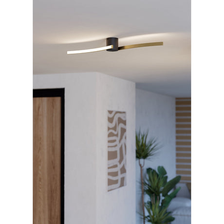 Aieta LED Flush Ceiling Light in black and brushed brass, with curved arms and illuminated white diffuser, fitted in a hallway ceiling near a plant and framed wall art.