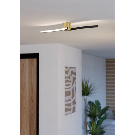 Aieta LED flush ceiling light with brushed brass cylindrical canopy and curved black and white LED arms, shown mounted on a hallway ceiling above a wall art and plant.
