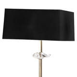 The Akira 3 Light Floor Lamp features an antique brass finish, a slim metallic stand, and a black fabric shade, with a decorative clear glass element shaped like a fish near the base of the shade.