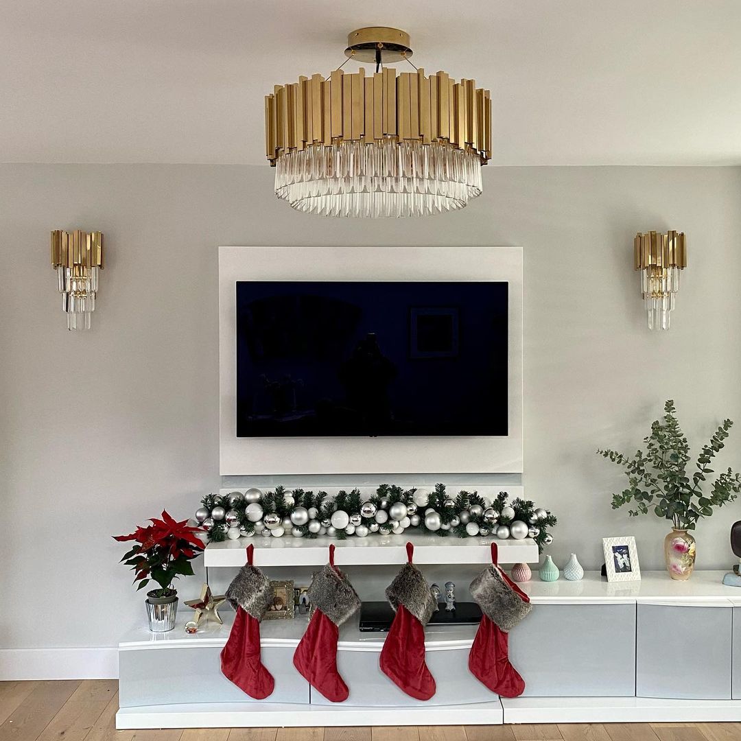 A modern living room with Art Deco influence highlights a wall-mounted TV and an elegant chandelier overhead. Beneath the TV, a garland adorned with holly enhances the gold finishes of the console, along with four red stockings. A poinsettia and framed photos sit nearby, while the Royal 2 Light Wall Light in Gold adds an extra touch of sophistication.