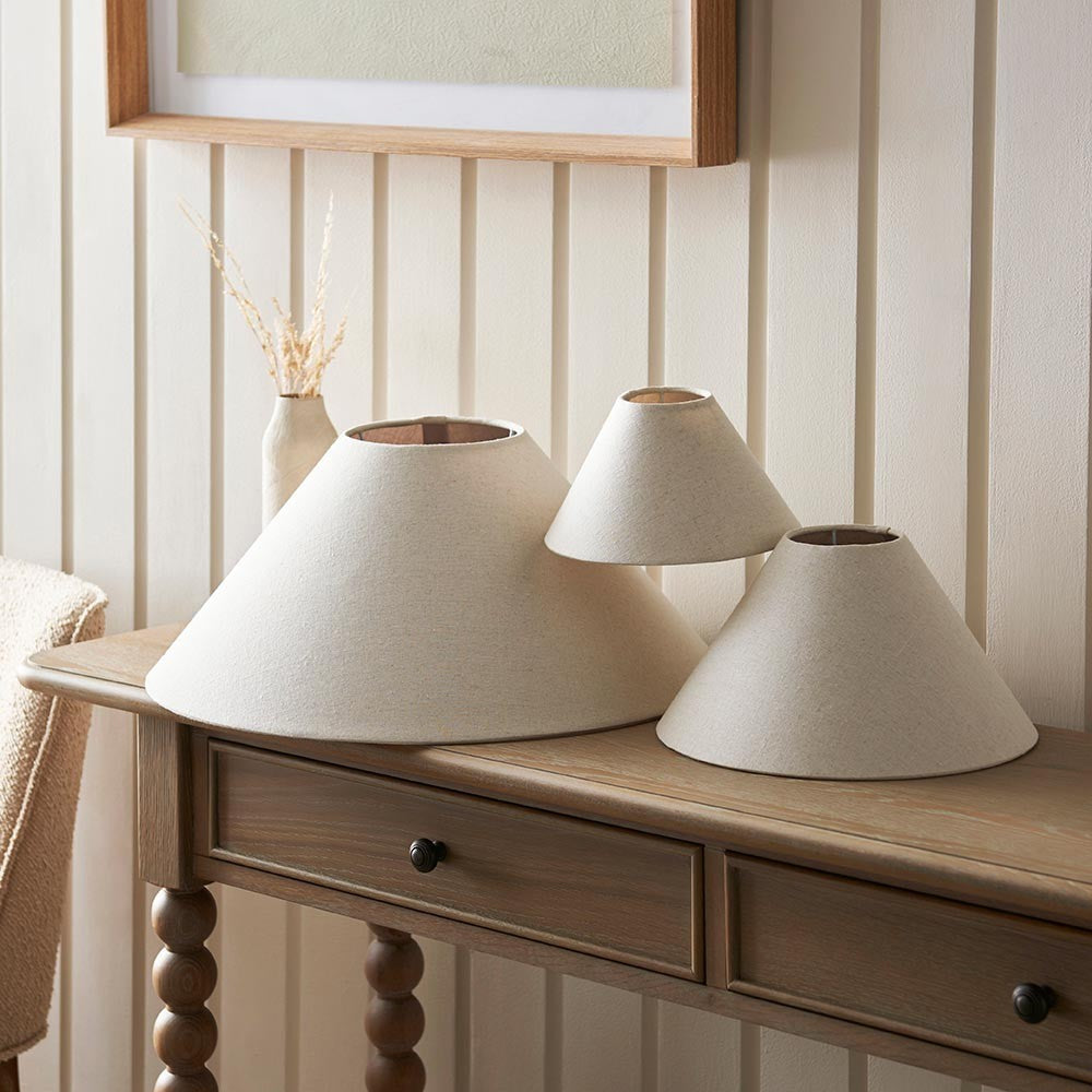 Aldmere tapered lamp shades in natural fabric, shown in three sizes on a wooden console table beneath a wall mirror.
