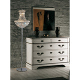 A vintage white dresser with ornate handles stands against a gray stone wall. The Alexandra 6 Light Crystal Floor Lamp in polished chrome to the left casts ambient light, highlighting two dark vases and an abstract painting atop the dresser.