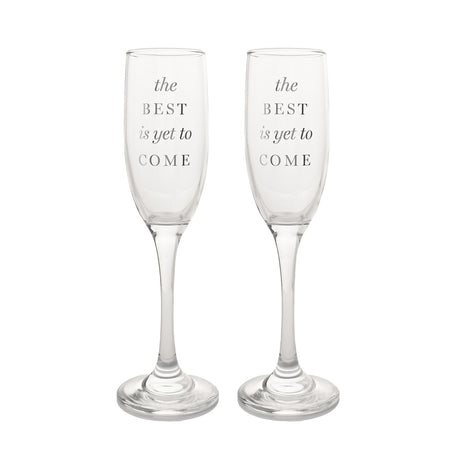 Set of two clear glass champagne flutes with tall slim bowls and slender stems, each printed with "the BEST is yet to COME" in silver-grey lettering, on a plain white background.