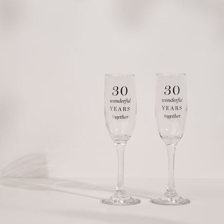 Pair of clear glass champagne flutes with tall narrow bowls printed "30 wonderful YEARS together" in black serif and italic text, set on a plain light background.