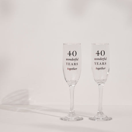 Set of two clear glass champagne flutes with tall narrow bowls and slim stems, printed "40 wonderful YEARS together" in black and red lettering, on a plain white background.