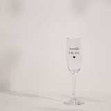 Clear glass champagne flute with a tall, slender bowl printed "beautiful BRIDE" in silver script and capitals above a small heart, shown on a plain light background.