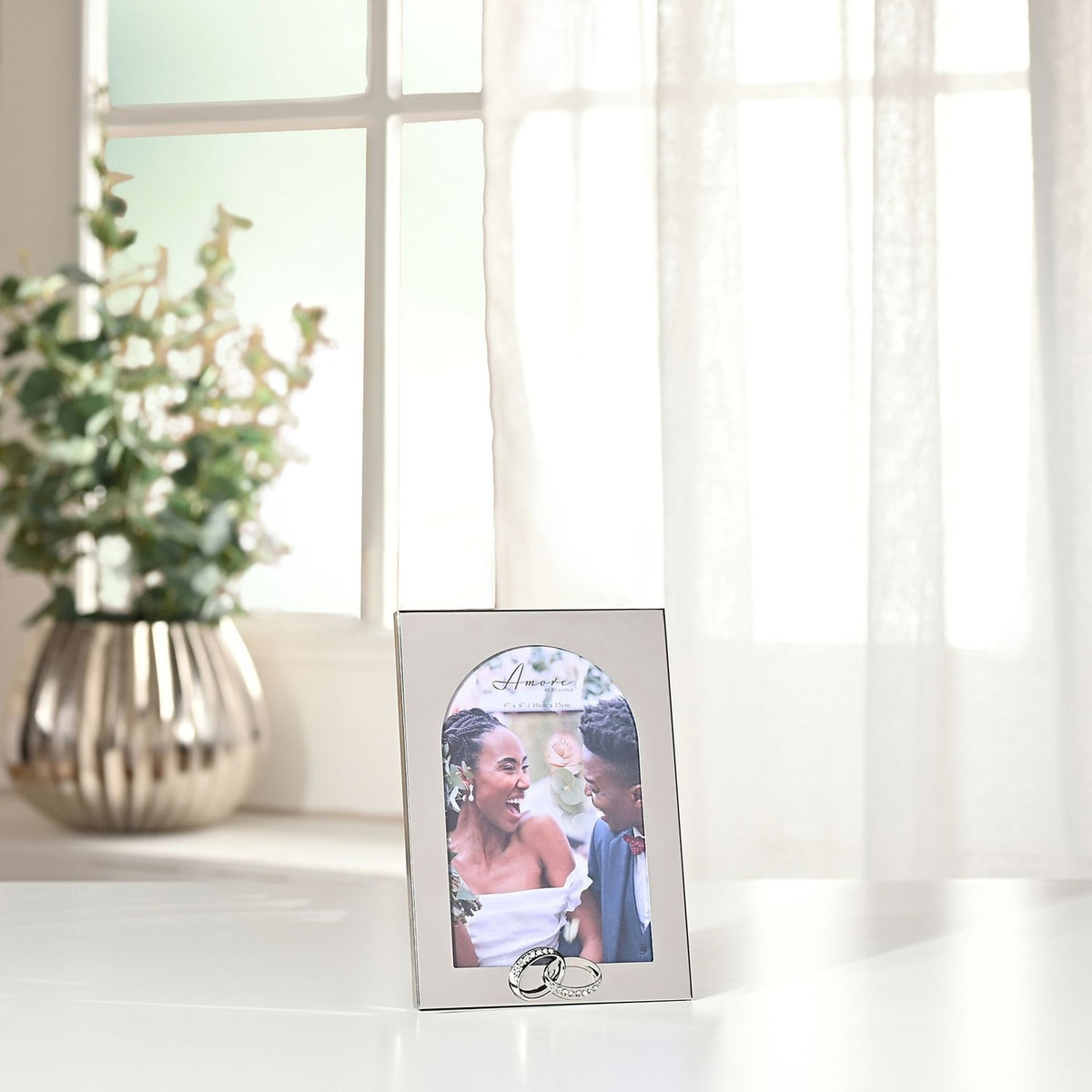 Arch Metal Photo Frame with Linked Rings 6 x 4in in shiny silver, displaying a wedding photo on a white surface near a window with sheer curtains and a potted plant.
