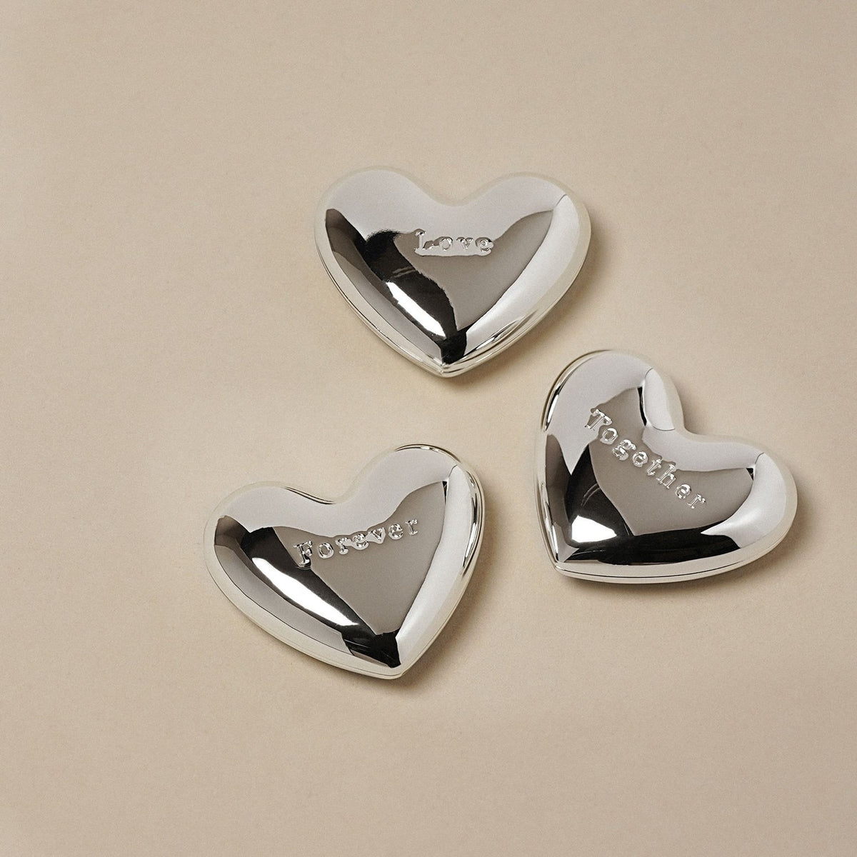 Three silverplated heart charm ornaments with a mirror-bright finish, each engraved with "Love," "Forever," or "Together," arranged on a plain light background.